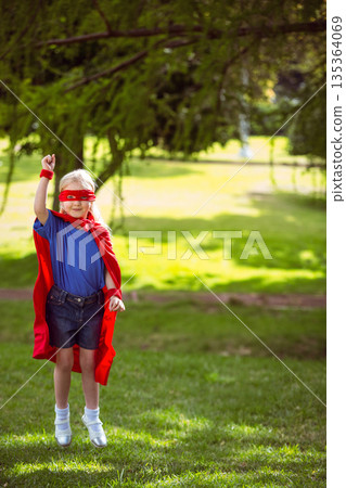 Girl is standing on sunlit park lawn under tree, wearing red superhero cape and mask 135364069