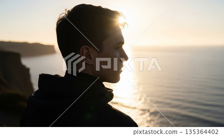 Young Asian woman in profile silhouette watching golden sunset over ocean water from coastal viewpoint wearing dark jacket 135364402