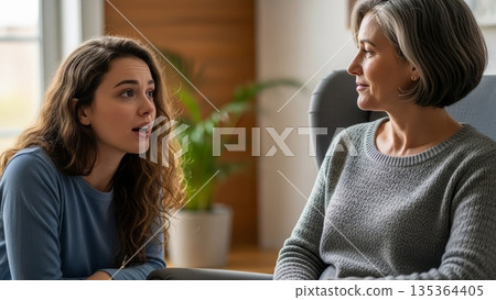 Young caucasian teenage daughter with curly hair having serious conversation with middle-aged blonde mother on living room sofa 135364405