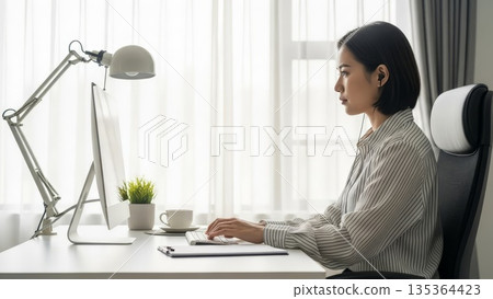 Asian businesswoman working on laptop computer at modern office desk with task lamp and coffee cup during daytime 135364423