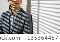 Professional asian businesswoman in dark suit standing by window with venetian blind shadows creating striped light pattern across business attire 135364457