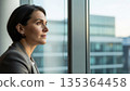 Professional Asian businesswoman in gray suit looking thoughtfully out office window with city skyline view during daytime meeting break 135364458