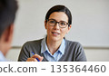 Professional Asian businesswoman with glasses smiling during office meeting consultation holding blue pen wearing grey cardigan 135364460