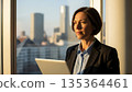 Professional Asian businesswoman with laptop looking out office window at city skyline during golden hour sunset 135364461