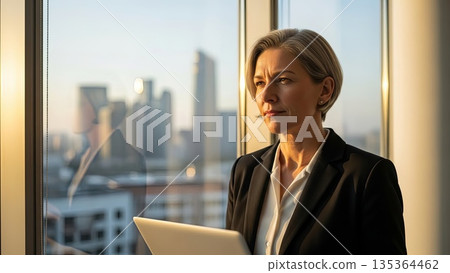 Professional asian businesswoman with laptop looking thoughtfully through office window at city skyline during golden hour 135364462