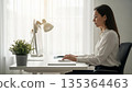 Professional Asian businesswoman working at modern office desk with computer keyboard and task lamp during daytime 135364463