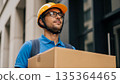 Professional Asian delivery worker wearing orange hard hat carrying cardboard package box looking up outdoors urban building background 135364465