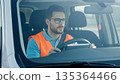 Professional Asian male delivery driver wearing orange safety vest and glasses driving white commercial van during daytime work shift 135364466