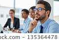 Professional bearded businessman with glasses thinking thoughtfully during corporate meeting with diverse colleagues in modern office boardroom 135364478