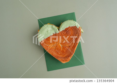 Heart shaped pistachio croissant on green background with copy space. Puff pastry trendy sweet dessert bakery. Creamy dough experiment Heart shaped pistachio croissant on green background with copy space. Puff pastry trendy sweet dessert bakery. Creamy dough experiment 135364904