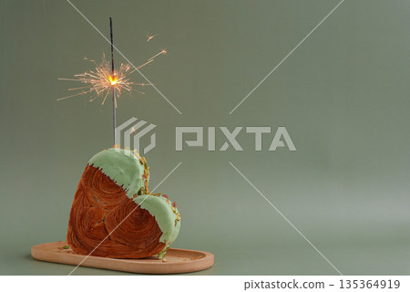 Festive holiday candle sparkler birthday anniversary celebration Heart shaped pistachio croissant on green background with copy space. Puff pastry trendy sweet dessert bakery. Creamy dough experiment  135364919