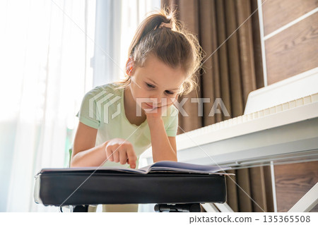 Young girl leaning on chair and reading sheet music before piano practice at home. Music study moment, preparation for lesson, concentration, and thoughtful childhood learning routine. Young girl leaning on chair and reading sheet music before piano practice at home. Music study moment, preparation for lesson, concentration, and thoughtful childhood learning routine. 135365508