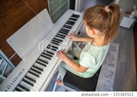 Little girl sitting at piano and playing music at home, expression and careful posture. Childhood music education, creative development, concentration, discipline, and calm learning atmosphere Little girl sitting at piano and playing music at home, expression and careful posture. Childhood music education, creative development, concentration, discipline, and calm learning atmosphere 135365521