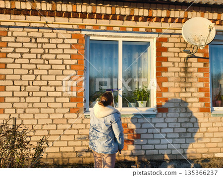 A woman stands outside and cleans a house window with a squeegee on a sunny day. A rear view of a woman cleaning a house window while standing outside. Ordinary household chores. A woman stands outside and cleans a house window with a squeegee on a sunny day. A rear view of a woman cleaning a house window while standing outside. Ordinary household chores. 135366237