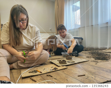 The family enjoys time together, sitting on the floor and creating small structures and objects out of cardboard and branches. The mother teaches her children creativity, developing their skills. 135366243