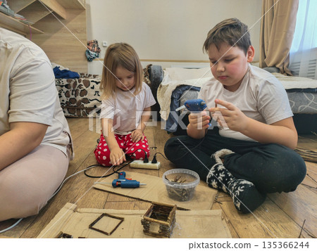 Children and mother enjoying creative crafting time together, building small structures with natural materials and hot glue guns, fostering imagination and family bonding. Children and mother enjoying creative crafting time together, building small structures with natural materials and hot glue guns, fostering imagination and family bonding. 135366244