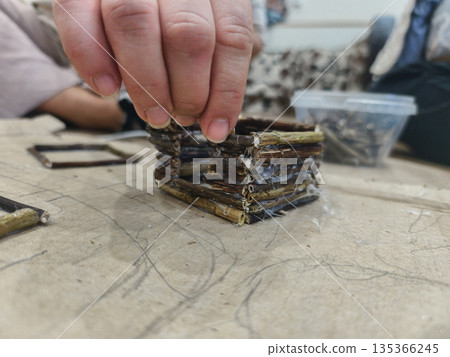 Person's hands carefully building a miniature house structure made from natural sticks, gluing them together on a work surface, emphasizing creativity and handmade projects. 135366245