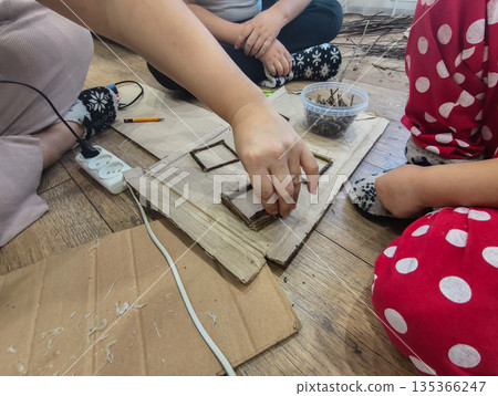 Mother and children creating a diy project, gluing twigs onto cardboard to build a house, fostering creativity and family bonding during an educational activity at home. Mother and children creating a diy project, gluing twigs onto cardboard to build a house, fostering creativity and family bonding during an educational activity at home. 135366247