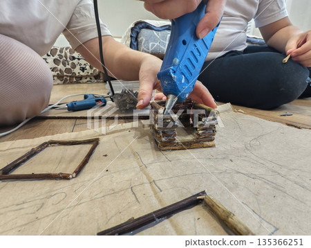 A family assembles a small wooden house out of twigs using a glue gun on the floor. Mother and child are engaged in creative crafting and learning together at home. A family assembles a small wooden house out of twigs using a glue gun on the floor. Mother and child are engaged in creative crafting and learning together at home. 135366251