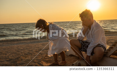 Two children, a boy and a girl, sitting and playing on a sandy beach during a beautiful sunset, sharing a quiet, pensive moment by the ocean as the sun dips below the horizon. Two children, a boy and a girl, sitting and playing on a sandy beach during a beautiful sunset, sharing a quiet, pensive moment by the ocean as the sun dips below the horizon. 135366311