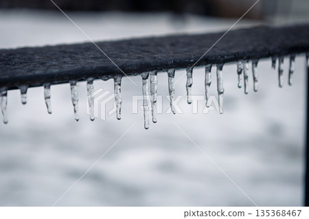 A metal fence is covered with frost, ice, and hanging icicles in a snowy landscape 135368467