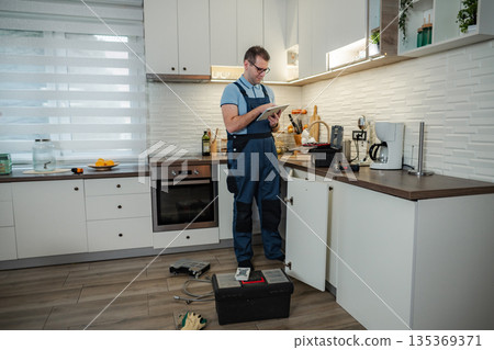 Plumber in overalls using tablet while repairing kitchen sink Plumber in overalls using tablet while repairing kitchen sink 135369371