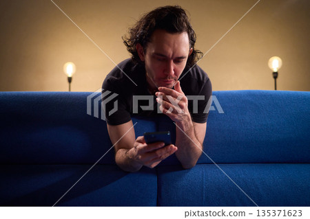 Chilled man on sofa, Casual individual reclining on sofa engrossed with mobile device, Unwinding man sits comfortably on sofa focused on his smartphone amid cozy modern decor Chilled man on sofa, Casual individual reclining on sofa engrossed with mobile device, Unwinding man sits comfortably on sofa focused on his smartphone amid cozy modern decor 135371623