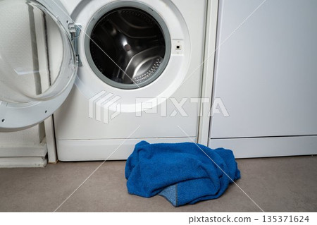 Blue towel lying on the floor next to an open washing machine. Everyday domestic scene showing laundry routine and household activity. Concept of home, cleaning, domestic, lifestyle, chores. Blue towel lying on the floor next to an open washing machine. Everyday domestic scene showing laundry routine and household activity. Concept of home, cleaning, domestic, lifestyle, chores. 135371624