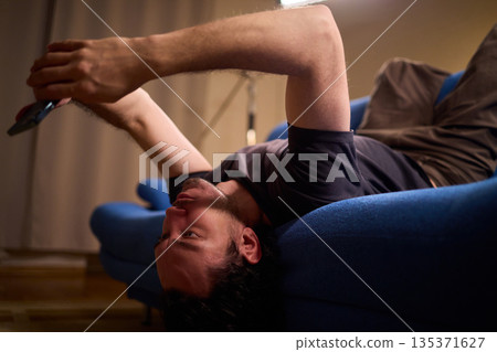 Chilled man on sofa, Casual individual reclining on sofa engrossed with mobile device, Unwinding man sits comfortably on sofa focused on his smartphone amid cozy modern decor Chilled man on sofa, Casual individual reclining on sofa engrossed with mobile device, Unwinding man sits comfortably on sofa focused on his smartphone amid cozy modern decor 135371627