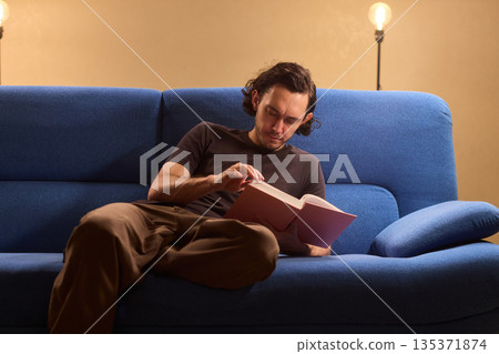 Individual sitting comfortably reading hardcover novel, Person lounges casually on soft blue sofa deeply engaged in hardcover novel during peaceful evening in urban apartment Individual sitting comfortably reading hardcover novel, Person lounges casually on soft blue sofa deeply engaged in hardcover novel during peaceful evening in urban apartment 135371874