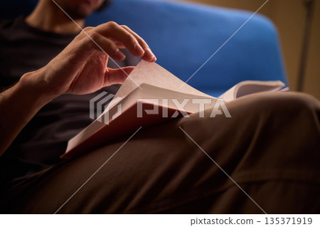Gentle scene with warm illumination, Person peacefully flipping pages under soft ambient light at home, Calm person immersed in book with cozy sweater and warm lamp glow 135371919