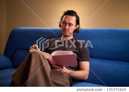 Man reads relaxingly, Relaxed man flipping through book, Individual leisurely reads on comfortable sofa, Person enjoying quiet evening with book and soft lighting 135371920