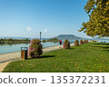 Peaceful lakeside promenade along Lake Balaton with flowering bushes, calm water, green park lawn, and distant hills creating a relaxing summer travel scene in Hungary 135372231