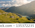 Scenic alpine mountain valley road leading toward the Pasterze Glacier with winding curves, remote mountain huts, and a turquoise glacial lake surrounded by green slopes and dramatic high Alpine peaks 135372235
