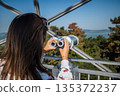 Woman looking through binoculars at Xantos viewpoint in Balatonboglar with panoramic views of Lake Balaton, forested hills, metal observation structure, and clear blue sky in Hungary 135372237