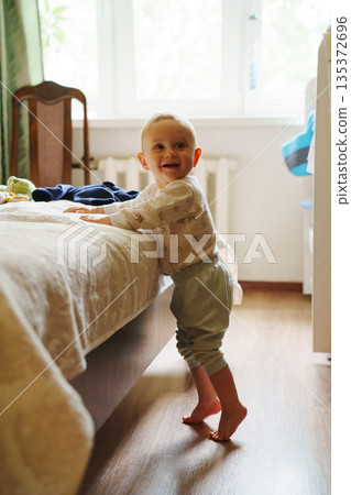 Happy toddler standing by the bed in a bright room Happy toddler standing by the bed in a bright room 135372696