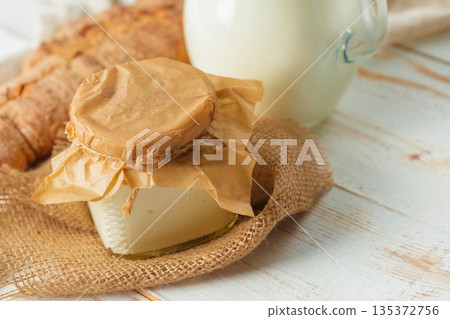 Milk and cheese sit on a wooden table with bread nearby in a rustic kitchen setting Milk and cheese sit on a wooden table with bread nearby in a rustic kitchen setting 135372756