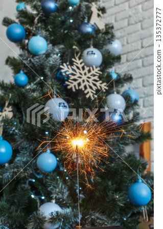 Sparkler shines in front of decorated Christmas tree with blue ornaments in indoor setting Sparkler shines in front of decorated Christmas tree with blue ornaments in indoor setting 135372777