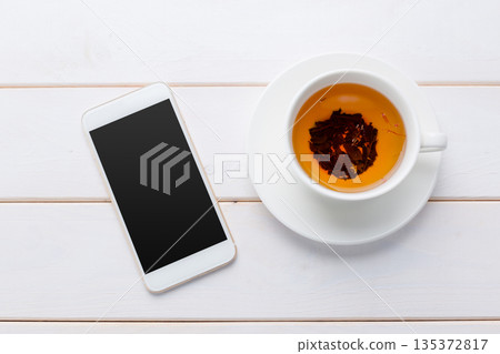 Warm cup of tea next to a smartphone on a wooden table surface in a cozy setting Warm cup of tea next to a smartphone on a wooden table surface in a cozy setting 135372817