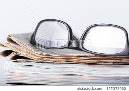 Reading glasses placed on a stack of newspapers and magazines in a simple setting 135372960