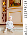 Child in a white dress sitting by religious artwork 135373013