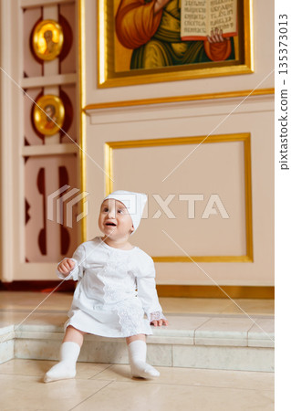 Child in a white dress sitting by religious artwork 135373013