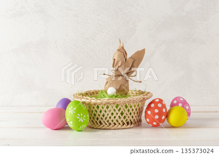 Colorful eggs and bunny wrapped in paper sit in a basket on a bright table surface 135373024