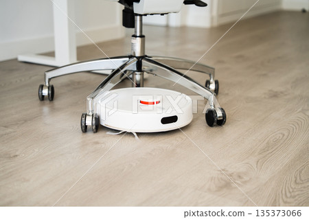 Smart robot vacuum cleaner working under office chair on wooden floor, automated cleaning device for modern home and workspace Smart robot vacuum cleaner working under office chair on wooden floor, automated cleaning device for modern home and workspace 135373066