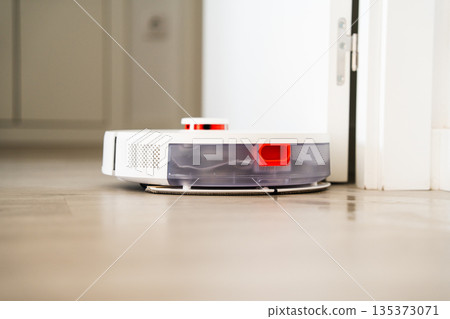Smart robot vacuum cleaner navigating doorway on wooden floor in modern apartment, autonomous home cleaning technology concept 135373071