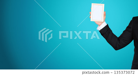 Person holds a blank frame in front of a blue background during an indoor event or activity 135373072