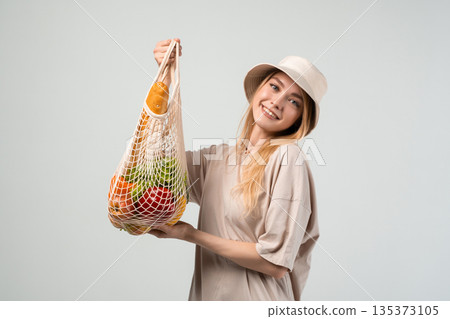 Smiling young woman in beige outfit and bucket hat holding reusable mesh grocery bag with fresh vegetables and bread 135373105