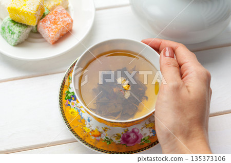 Hand holding cup with tea beside colorful sweets on table at indoor location in afternoon Hand holding cup with tea beside colorful sweets on table at indoor location in afternoon 135373106