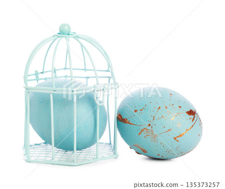 Decorative blue egg placed next to a small bird cage on a white surface Decorative blue egg placed next to a small bird cage on a white surface 135373257