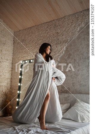 A woman with black hair wakes up in the morning in a white blanket and catches the rays of the sun. The portrait is creative, with brown eyes. High quality photo 135374107